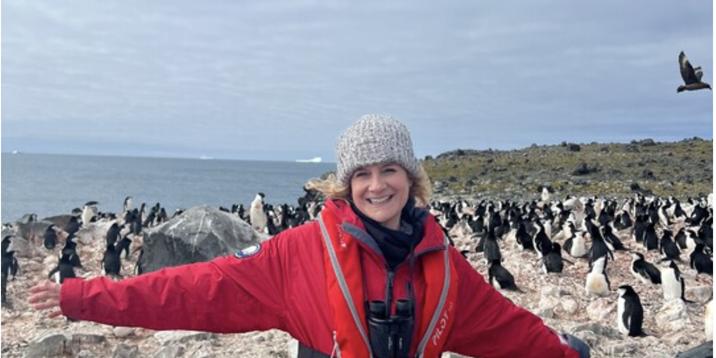 woman in red jacket and hat with her arms out, and penguins behind her. Photo from Penguin Island, Southern Ocean (January 1, 2024)