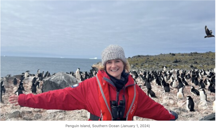woman in red jacket and hat with her arms out, and penguins behind her. Photo from Penguin Island, Southern Ocean (January 1, 2024)