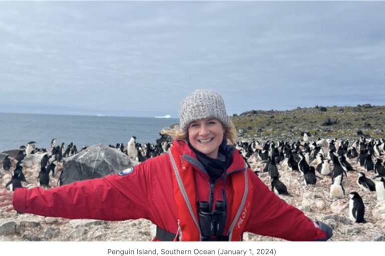 woman in red jacket and hat with her arms out, and penguins behind her. Photo from Penguin Island, Southern Ocean (January 1, 2024)