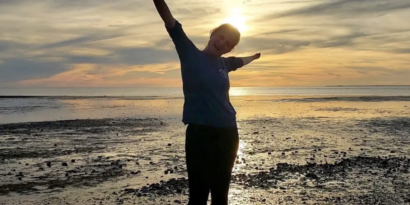 ID: Woman standing and extending her arms on a beach with a beautiful sunset in the background.