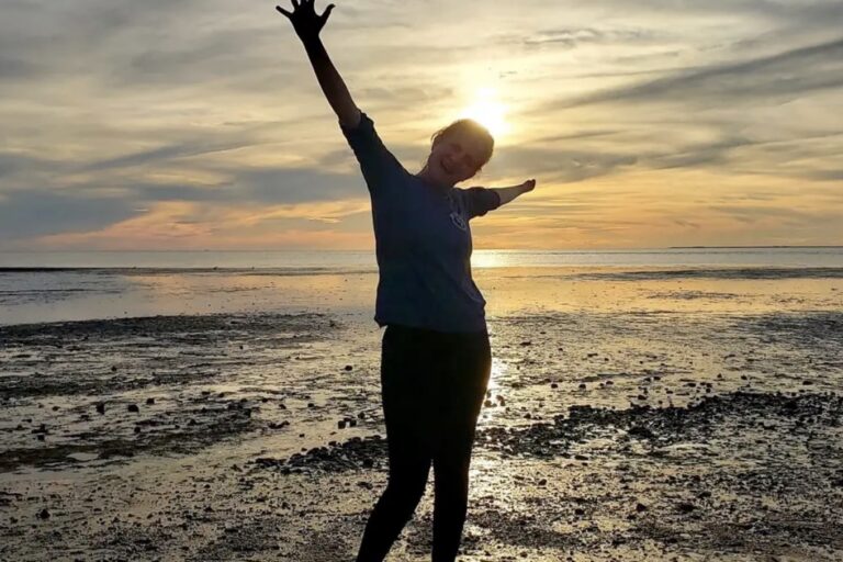 ID: Woman standing and extending her arms on a beach with a beautiful sunset in the background.