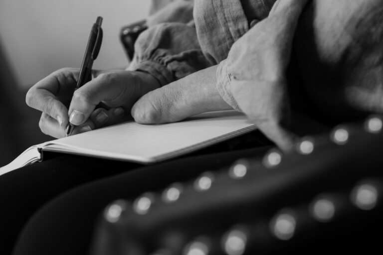 ID: Image of a person writing in a notebook, focusing on their hands and pen. One hand is limb different.