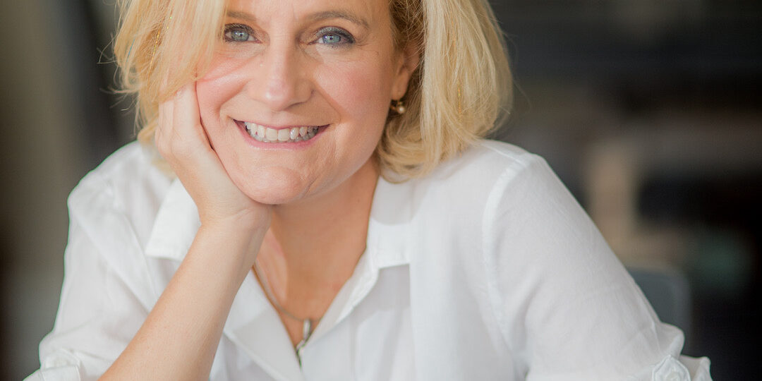 White female, smiling, wearing a white shirt; one hand resting on her chin, limb different hand resting on computer.