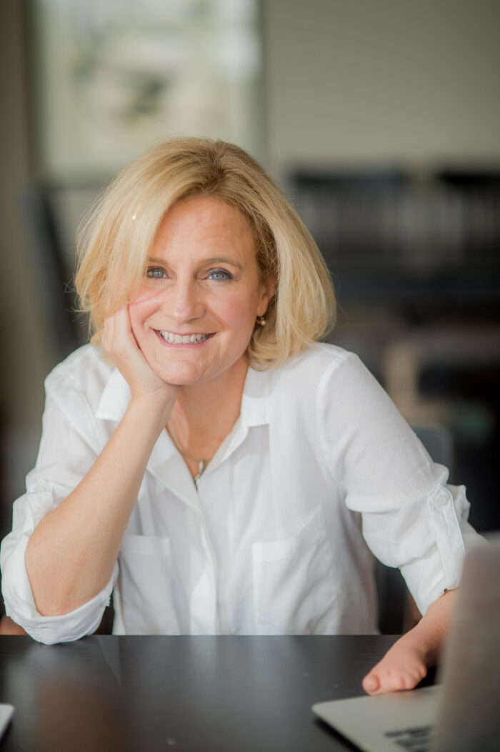 White female, smiling, wearing a white shirt; one hand resting on her chin, limb different hand resting on computer.