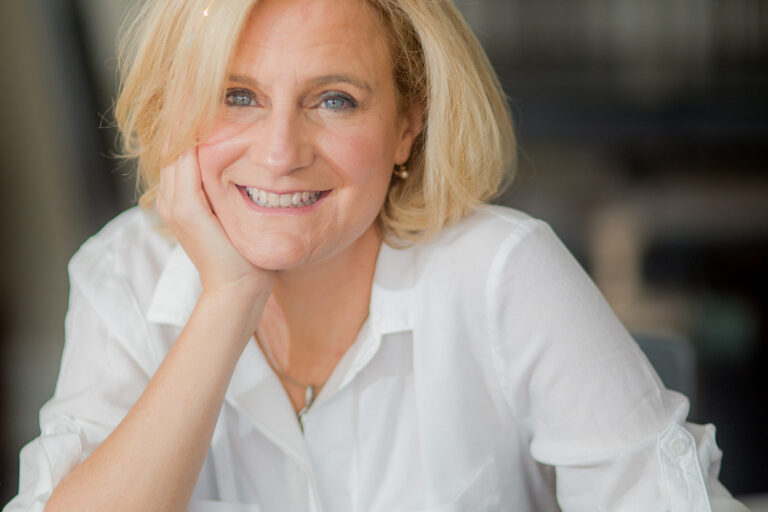 White female, smiling, wearing a white shirt; one hand resting on her chin, limb different hand resting on computer.