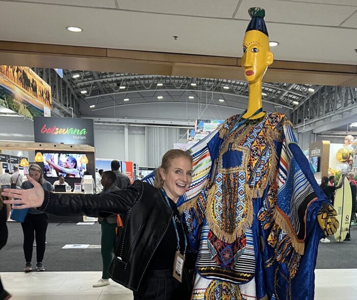 ID: Me (white female) posing with arms outstretched next to a tall, colorful, stylized figure in traditional African attire. At WTM Africa
