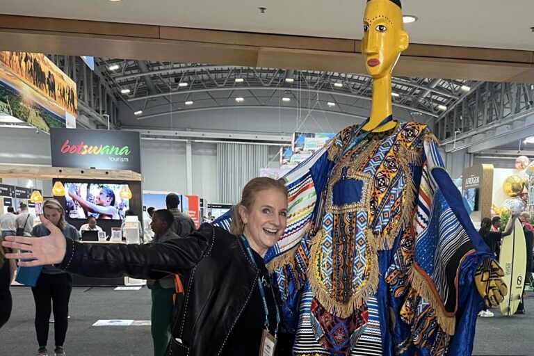 ID: Me (white female) posing with arms outstretched next to a tall, colorful, stylized figure in traditional African attire. At WTM Africa