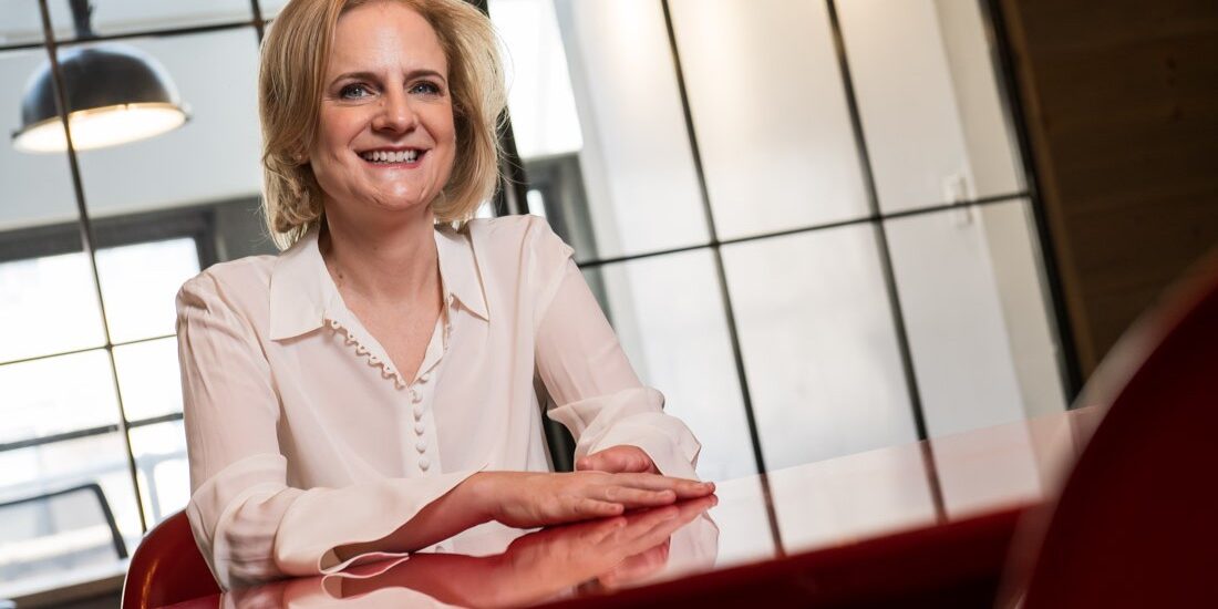 newsletter june 2026 Speaker and Author Ruth Rathblott seated at a red table in a bright workspace, smiling, with her limb difference visible. Photo: John DeMato