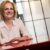 Speaker and Author Ruth Rathblott seated at a red table in a bright workspace, smiling, with her limb difference visible. Photo: John DeMato
