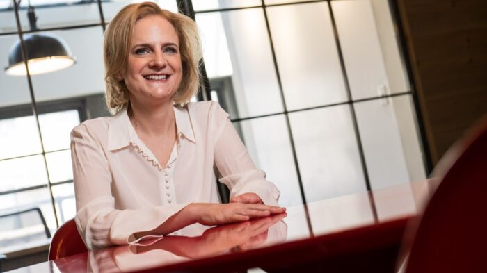 newsletter june 2026 Speaker and Author Ruth Rathblott seated at a red table in a bright workspace, smiling, with her limb difference visible. Photo: John DeMato
