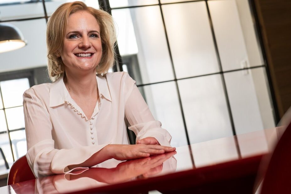 Speaker and Author Ruth Rathblott seated at a red table in a bright workspace, smiling, with her limb difference visible. Photo: John DeMato